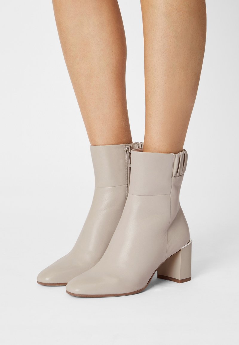 Bottines beiges à talons carrés, avec fermetures éclair latérales et détails froncés en haut, portées sur des jambes nues sur un fond uni.