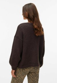 Przewymiarowany ciemnobrązowy sweter o teksturowanym wzorze dzianiny, z opuszczanymi ramionami i ściągaczem na dole, zestawiony z pasiastymi zielono-brązowymi spodniami.