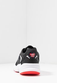 Černá atletická bota s texturovaným koženým svrškem, bílými akcenty a červenobílým logem na zadní části. Silná bílá mezipodešev s červenou podrážkou.