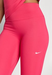 Růžové sportovní legíny vyrobené z hladkého, elastického materiálu. Mají vysoký pas a bílé logo Nike na dolní části nohy.