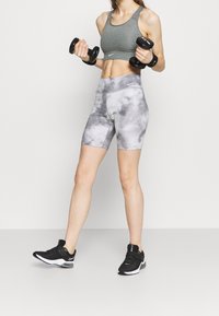 Šedá sportovní podprsenka a šedé šortky s vysokým pasem a mramorovým vzorem. Černé sportovní boty s bílými doplňky. Drží černé činky.