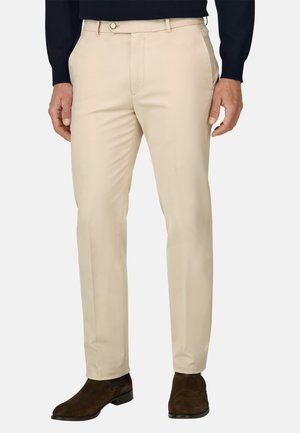 Homme portant un pantalon beige slim, des bottines en daim marron foncé et un haut noir à manches longues, debout les mains relâchées le long du corps.