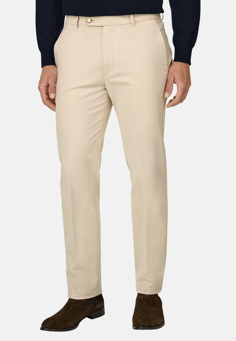 Homme portant un pantalon beige slim, des bottines en daim marron foncé et un haut noir à manches longues, debout les mains relâchées le long du corps.