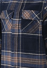 Chemise en flanelle à carreaux bleu marine et marron, avec deux poches poitrine à boutonnière, une fermeture à boutons sur le devant et un tissu texturé.