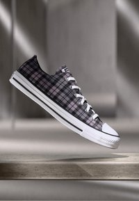 Sneaker a metà altezza a scacchi neri e viola con lacci bianchi e punta in gomma, sospeso sopra una superficie di pietra chiara.
