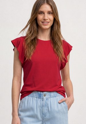 Młoda kobieta z długimi jasnobrązowymi włosami, ubrana w czerwoną, falbanianą, bezrękawnikową bluzkę i jasnoniebieskie spodnie z elastycznym pasem, uśmiechająca się z ręką w kieszeni.