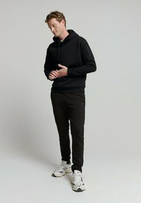 Zwarte hoodie met een voorzak en trekkoord, gecombineerd met zwarte joggers. De joggers hebben een subtiele logo en een fijne textuur.