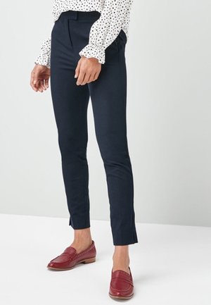 Femme portant un pantalon slim bleu marine longueur cheville, un chemisier blanc à pois noirs et des mocassins en cuir rouges, debout devant un fond uni.