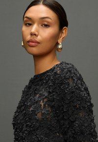 Zwarte bloemenkant top met een textuurafwerking, voorzien van 3D-stoffen bloemen. Gouden hoopsieraden bieden een contrast tegen de donkere outfit.