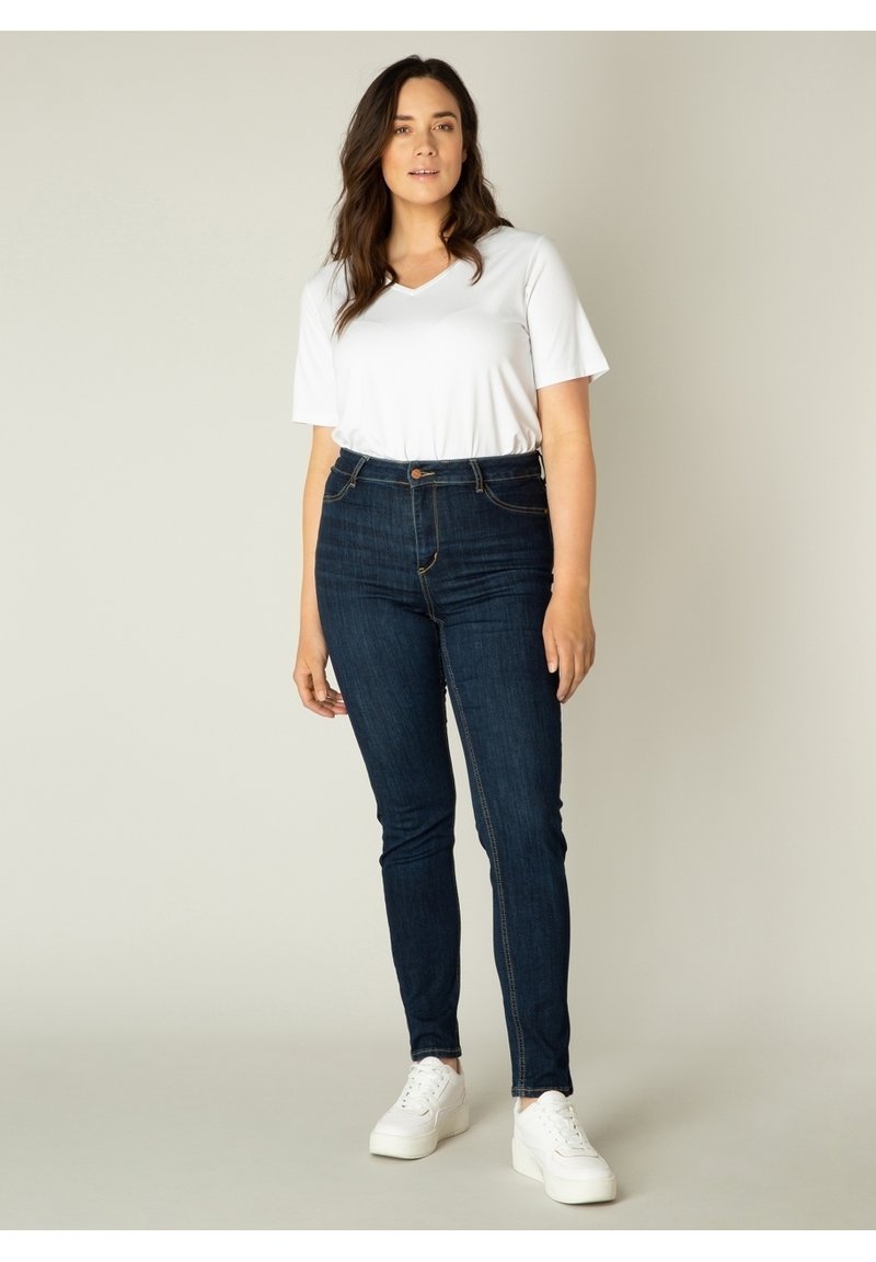 Donkerblauwe high-waisted jeans met een slim-fit ontwerp, gedetailleerde stiksels en een knoopsluiting, gecombineerd met een wit T-shirt met korte mouwen en V-hals, en witte sneakers.