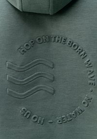 Zelená mikina s textúrovaným kruhovým dizajnom a nápisom "HOP ON THE BORN WAVE" a zrelaxovane vlnité línie vyrazené.