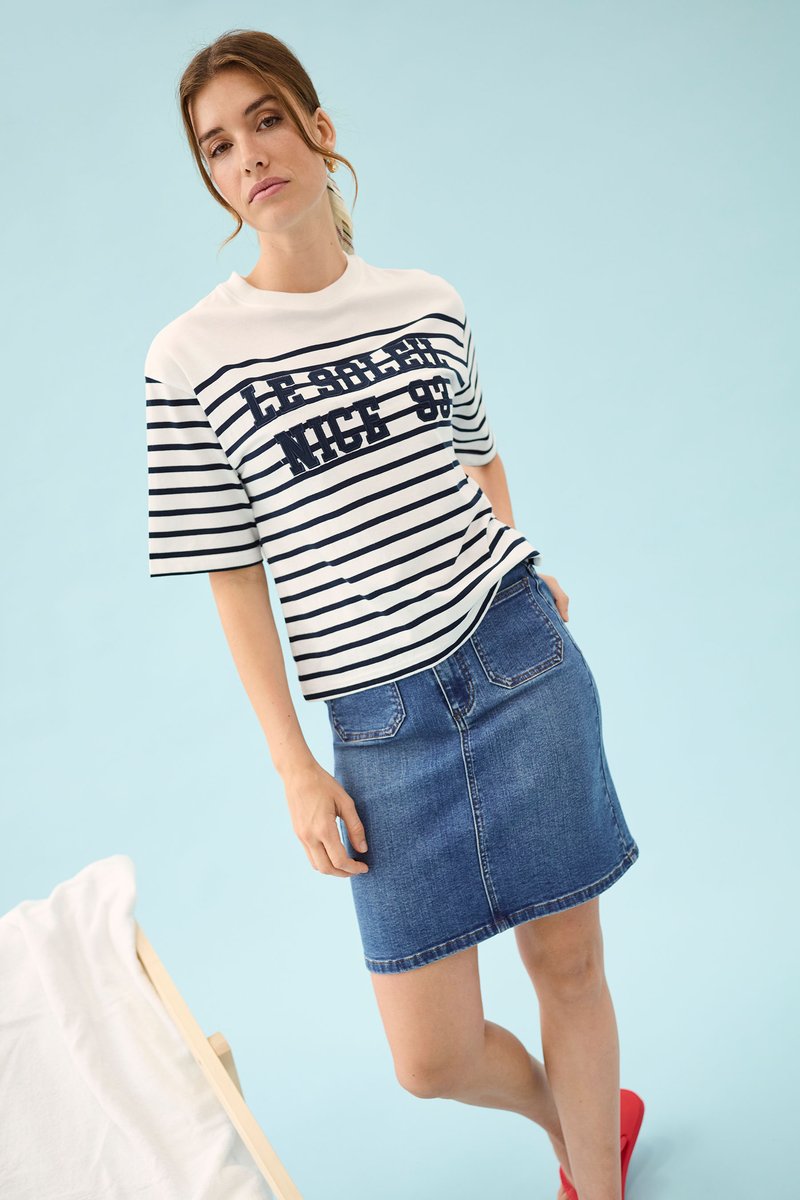 Jeune femme portant un t-shirt rayé avec l'inscription « Le Soleil Nice 89 », jupe en jean et chaussures rouges, debout sur un fond bleu clair.