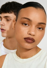 Zwei Models sind abgebildet, eines mit dunklem Haar und goldenen Kettenhalsketten. Das Model im Vordergrund trägt ein weißes Oberteil, hat ein natürliches Makeup und volle Lippen.