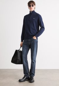 Pulover cu fermoar bleumarin, blugi din denim, pantofi din piele neagră și o geantă texturată neagră. Modelul stă în fața unui fundal deschis.