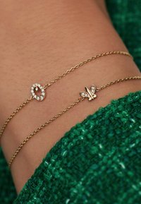 Kullanılmış altın zincir bilezik, şeffaf taşlarla süslenmiş dairesel bir aksanı ve bir kelebek charm'ı içeriyor, dokulu yeşil bir arka planda sergileniyor.