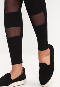 Leggings noirs avec des panneaux opaques et transparents, associés à des baskets noires à enfiler avec une semelle blanche. Texture lisse sur l'ensemble.
