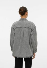 Giubbino oversize in denim grigio con colletto a punta, spalle scese e una piega centrale sul retro. Texture liscia, design semplice.