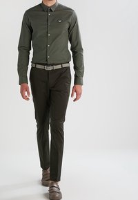 Olijfgroene overhemdbloes met een subtiel streepjespatroon, gecombineerd met donkere broeken en een lichtgrijze riem, heeft een klein logo op de borst.