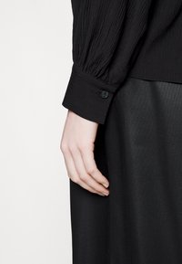 Main féminine reposant sur le côté, portant une chemise noire à manches longues texturée avec un poignet boutonné et une jupe noire à motif subtil.