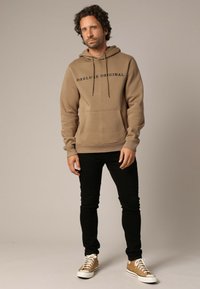 Man die staat in een bruine hoodie met de tekst "DEELUXE ORIGINAL", zwarte slim-fit broek en tan hoge sneakers met witte veters.