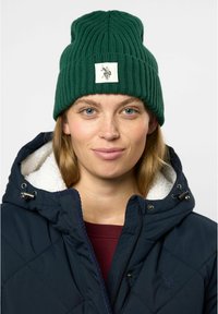 Grønn ribbestrikket lue med et firkantet merke med grafisk design. Båret med en marineblå quiltet jakke foret med hvit fleece.