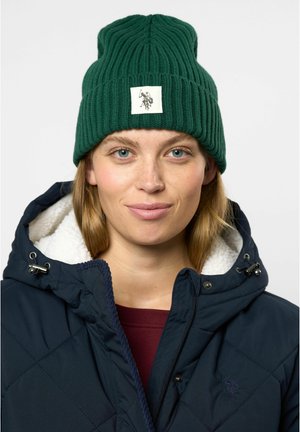 Green ribbed knit beanie with a square patch featuring a graphic design. Worn with a navy quilted jacket lined with white fleece.