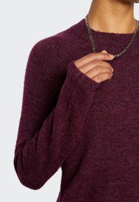 Pull en tricot bordeaux avec manches longues et coupe décontractée. Présente des poignets côtelés et un tissu doux et texturé. Collier chaîne visible.