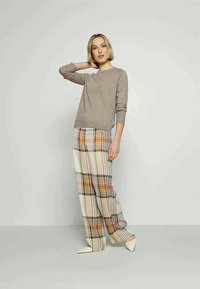 Gaiši brūns džemperis ar apaļu kakla izgriezumu apvienots ar krēmkrāsas un brūnu šaha raksta plašām biksēm; apģērbu noslēdz balti kurpju pirksti.