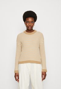 Maglione a righe beige e crema con scollo rotondo a costine e polsini, realizzato in materiale a maglia testurizzato, indossato con pantaloni a righe.
