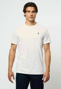 Balta trumpa rankovėmis marškinėlių pagaminta iš minkštos medžiagos, turinti mažą tamsų logotipą kairėje krūtinės pusėje; klasikinis apvalus kaklas ir tiesus apvadas.