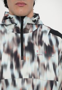 Pullover cu glugă cu un model abstract estompat în nuanțe de gri, albastru și portocaliu. Are un fermoar la jumătate în față, ajustări elastice pentru glugă și un logo roșu.