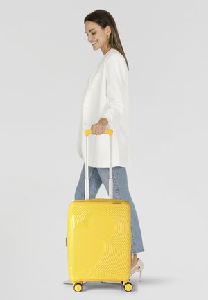 Valise rigide jaune avec un motif en spirale texturé et une silhouette de Mickey Mouse. Dispose d'une poignée télescopique et de quatre roues.