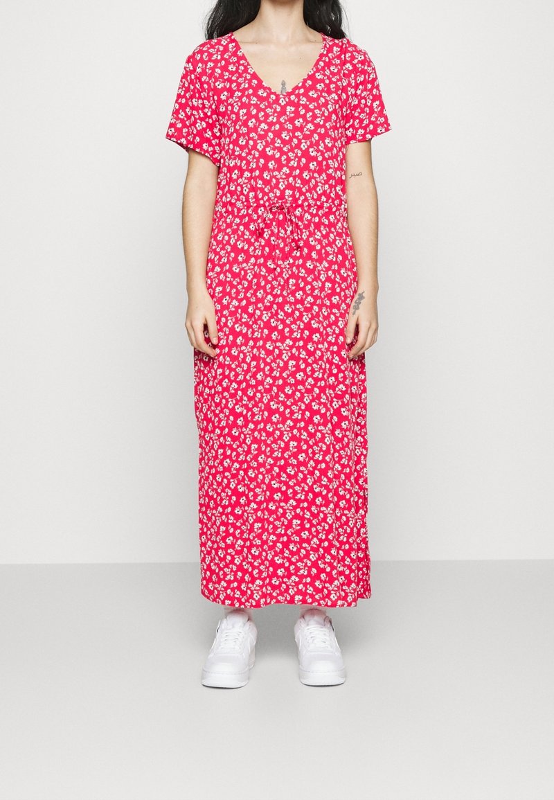 Femme portant une robe maxi rouge à fleurs avec des manches courtes et des baskets blanches, se tenant devant un fond clair.