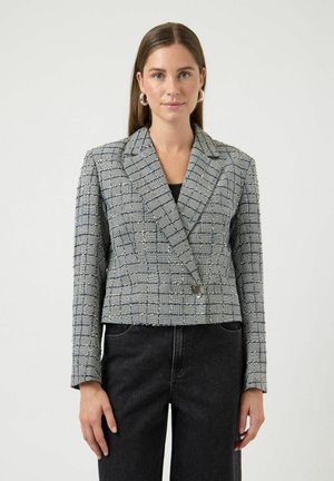 Vrouw met steil bruin haar, zilveren creolen, een grijze geruite blazer met één knoop en zwarte high-waisted jeans, staand tegen een witte achtergrond.