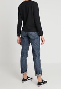 Felpa nera con collo rotondo, abbinata a jeans in denim blu con risvolti e sneakers nere con dettagli bianchi.