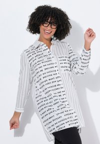 Camicia bianca con bottoni, caratterizzata da una combinazione di testo in grassetto e righe verticali. Include una tasca sul petto, maniche lunghe e una vestibilità rilassata.