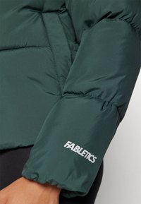 Zelená pufovaná bunda s rukávom, ktorý má zGatherovanú elastickú manžetu a výrazné biele logo "FABLETICS". Hladký a lesklý povrch.