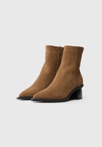 Bottes montantes en suédine marron avec un bout pointu et un talon bloc, dotées d'une fermeture éclair sur le côté et d'une texture lisse. Logo sur le côté.