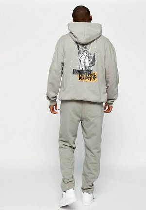 Persona que lleva puesta una sudadera con capucha y pantalones grises con un gráfico de la Estatua de la Libertad y texto en la parte trasera de la sudadera, de pie contra un fondo liso.