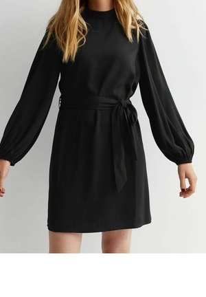 Femme portant une robe noire à manches longues avec une ceinture nouée à la taille, posant devant un fond gris clair uni.