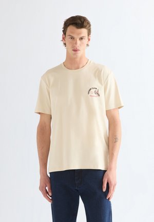 Jeune homme aux cheveux courts et bruns, portant un t-shirt beige avec l'impression "DENIM CLUB WRANGLER" et un jean bleu foncé, debout devant un fond blanc.