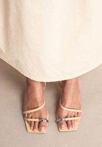 Sandales à talons blocs en cuir beige clair avec des brides fines et un accent argenté. Dessus carré et texture lisse.