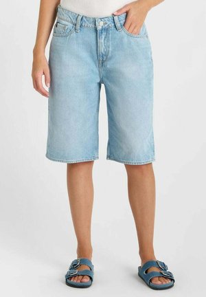 Pantaloncini di jeans azzurri alla lunghezza del ginocchio indossati da una persona in piedi, abbinati a sandali blu con doppie fibbie.