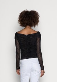 Top nero con design off-shoulder, tessuto arricciato e maniche trasparenti con pois. Indossato con pantaloni bianchi, che mostrano una silhouette aderente.
