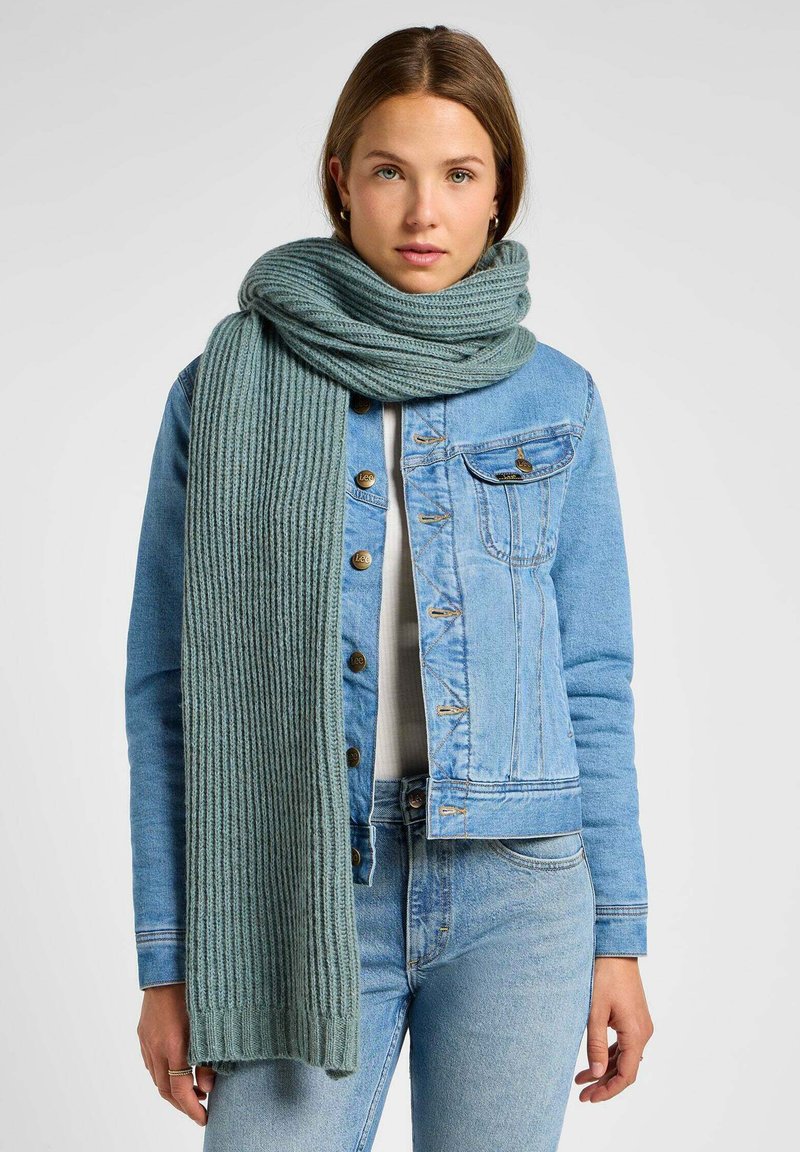 Jasnoniebieska dżinsowa kurtka w połączeniu z długim, prążkowanym, jasnoróżowym szalem. Szal ma miękką teksturę i strzępione krawędzie, oferując ciepło.