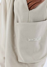Sac en tissu beige avec une texture diagonale, doté d'une poche latérale et d'un patch logo blanc bien visible portant l'inscription "PXP".