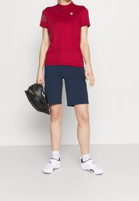 Červená košile s krátkým rukávem a jemným vzorem na rameni, doplněná námořnickými šortkami. Model drží černou helmu a má na sobě bílé boty.