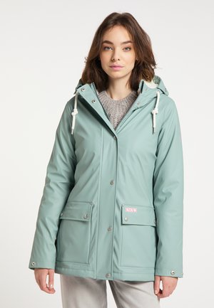 Giovane donna che indossa un impermeabile verde chiaro con cappuccio, tasche frontali e coulisse bianche, in piedi davanti a uno sfondo semplice.