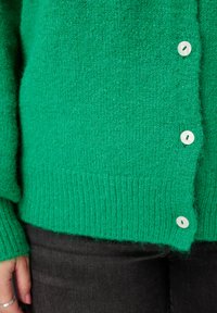 Groen gebreid vest met een geribde zoom en manchetten, voorzien van witte knopen. De textuur oogt zacht en pluizig, wat zorgt voor een gezellige uitstraling.