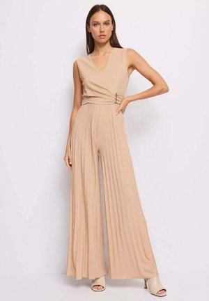 Donna che indossa una tuta beige senza maniche con scollo a V, pantaloni larghi plissettati e fibbia laterale, in piedi contro uno sfondo bianco.